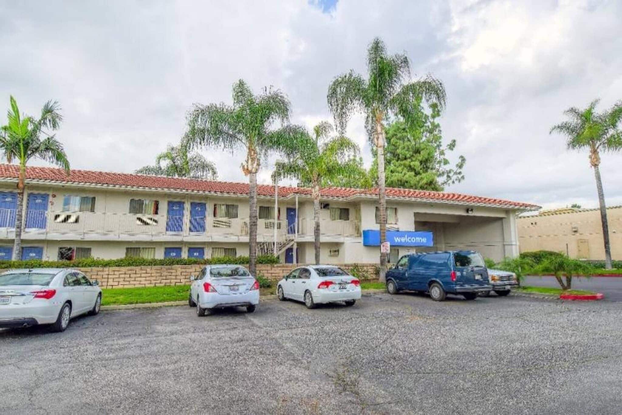 A multi-story building with a 'welcome' sign, a parking area with several cars, and palm trees.