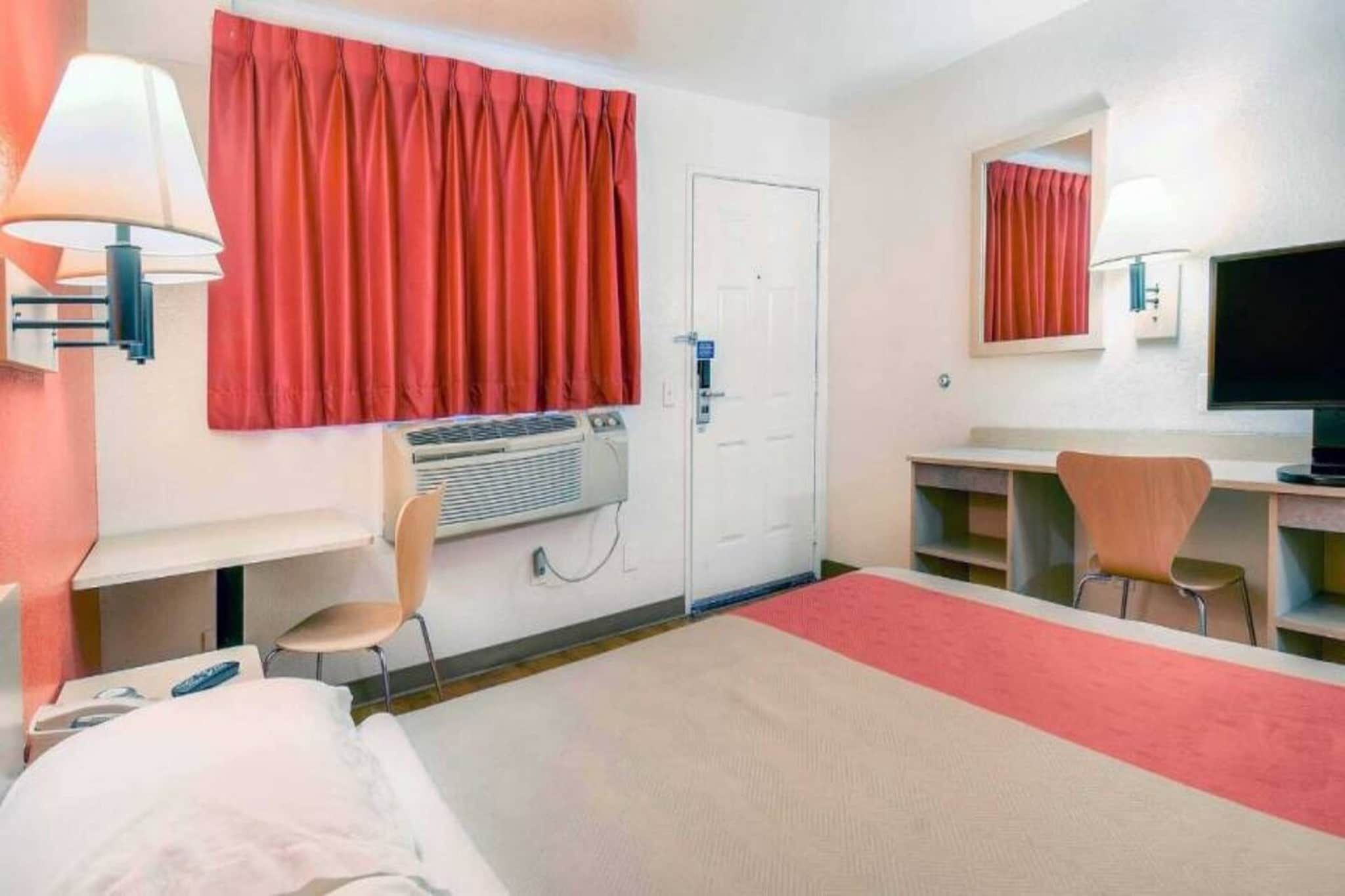 A hotel room with a bed, a desk, a chair, an air conditioning unit, a television, and a mirror.