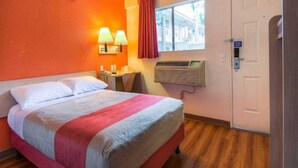 A hotel room with a bed, a desk, a chair, a window with curtains, an air conditioning unit, and a door. - 1 QUEEN BED | DELUXE (Chino)