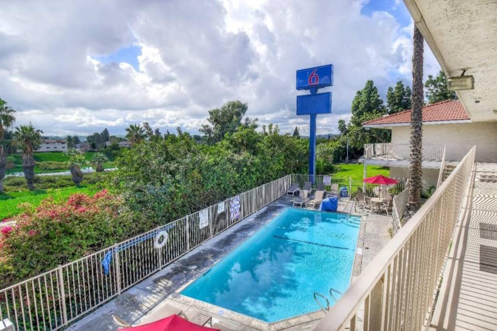 A swimming pool with a fenced area, a blue sign with the number 6, and a view of a green landscape with trees and a building in the background.