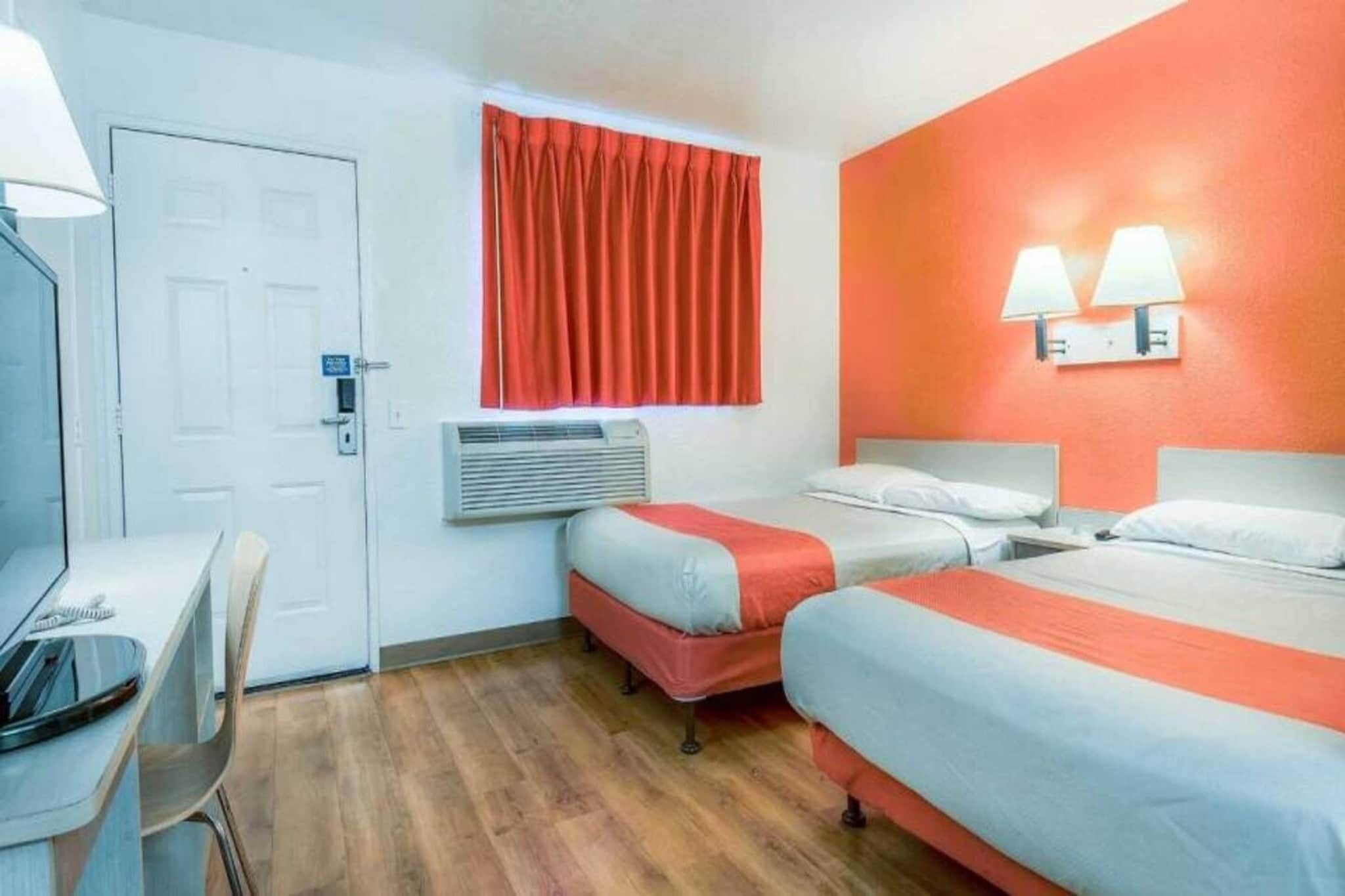A hotel room with two beds, a desk, a chair, a window with curtains, an air conditioning unit, and two wall-mounted lamps.
