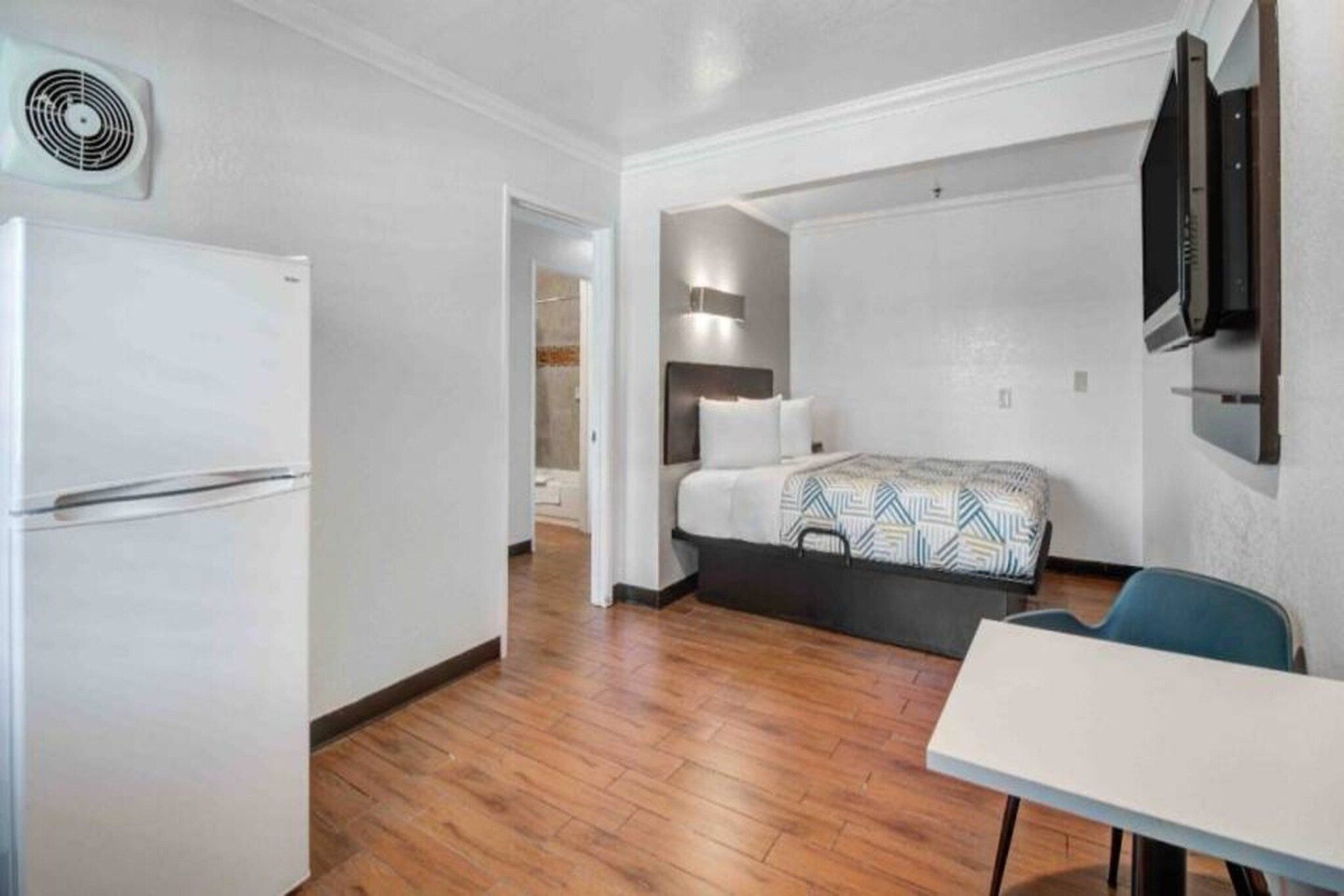 A compact hotel room with a bed, a small table, a chair, a TV mounted on the wall, and a ceiling fan.