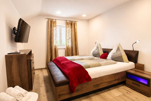 Ein modernes Schlafzimmer mit einem Holzbett, einem an der Wand montierten Fernseher, einem Nachttisch mit Lampe und einem Fenster mit Vorhängen.