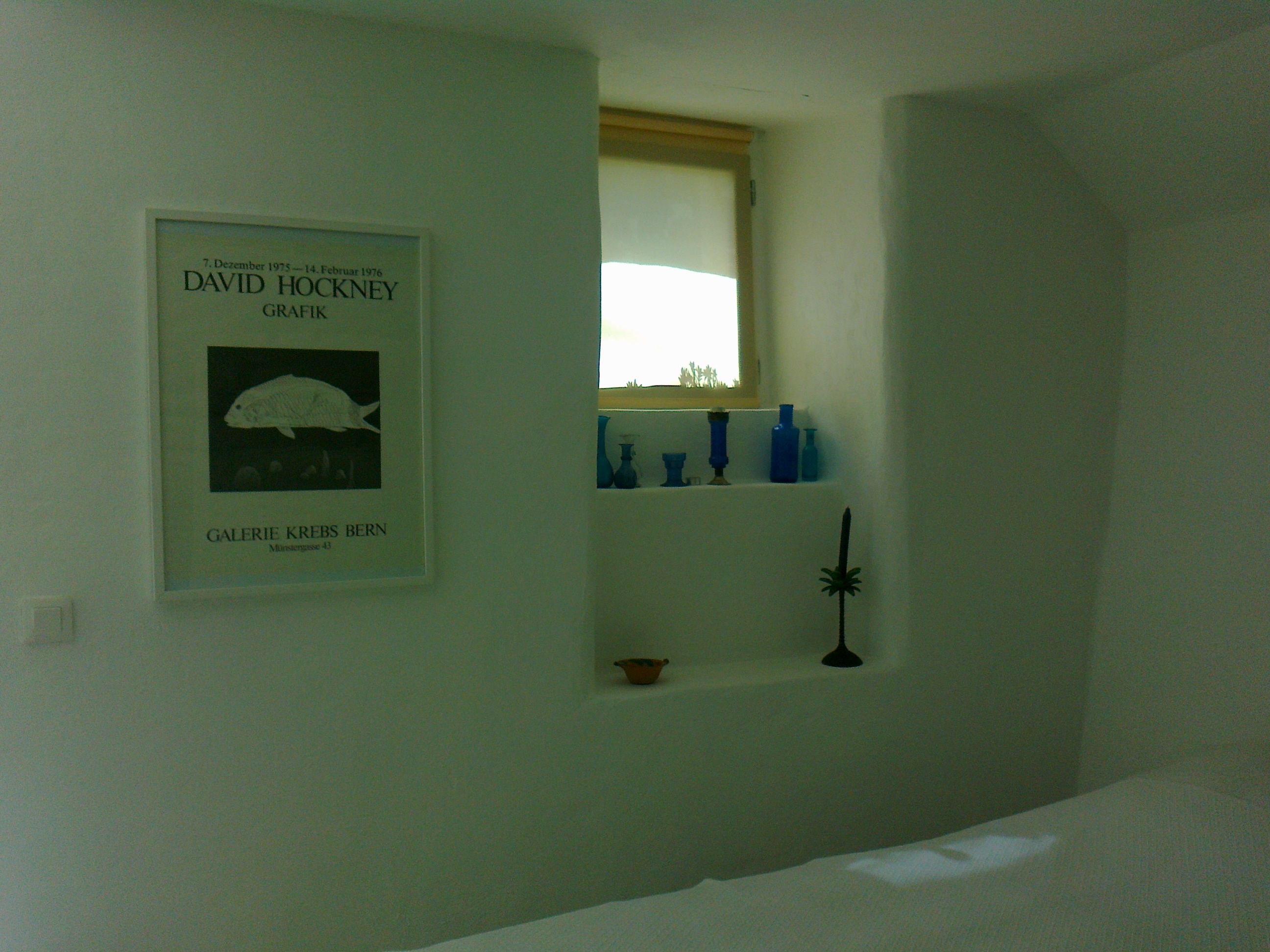 A room with a framed artwork on the wall, a window with a view, and a shelf with blue bottles.