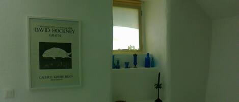 A room with a framed artwork on the wall, a window with a view, and a shelf with blue bottles.