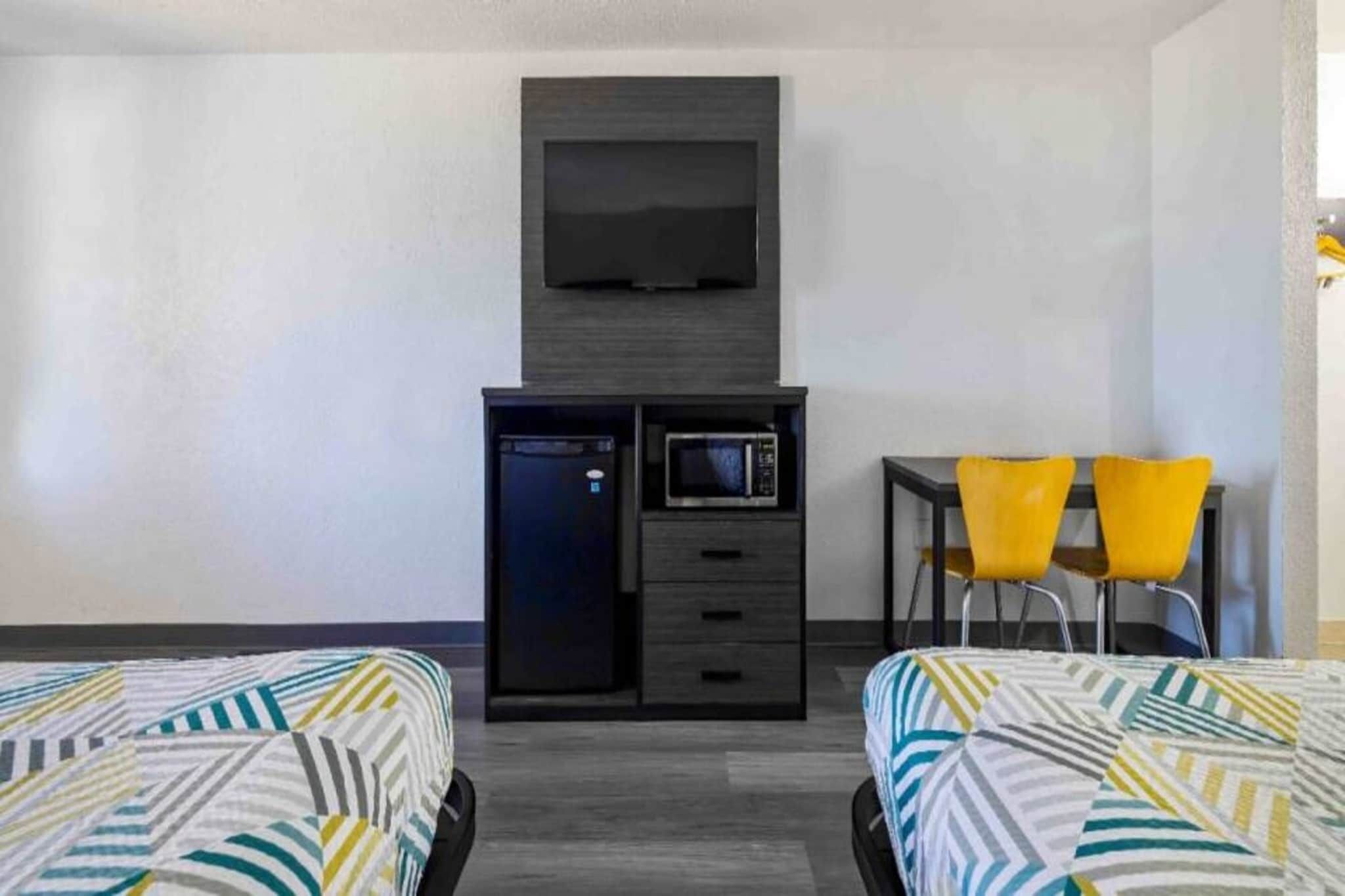 Ein Hotelzimmer mit zwei Betten, einem Schreibtisch, einer Mikrowelle, einem Fernseher und zwei gelben Stühlen.