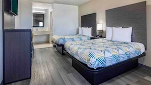 Quarto de hotel com duas camas, uma escrivaninha, uma televisão e um banheiro visível por uma porta aberta.