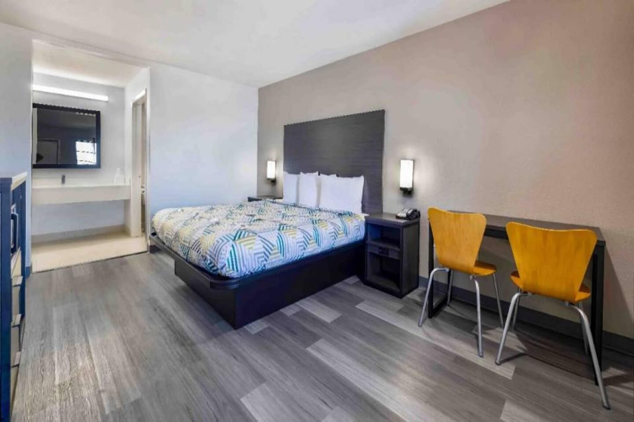 Una habitación de hotel con cama, mesita de noche, dos sillas y televisor montado en la pared.