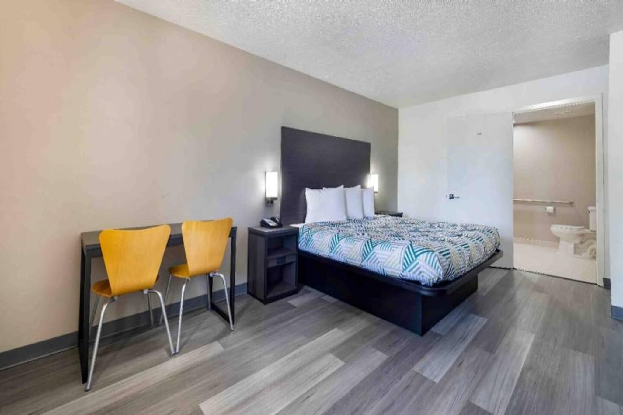Habitación de hotel con cama, dos sillas, una mesita de noche y un baño visible a través de una puerta abierta.