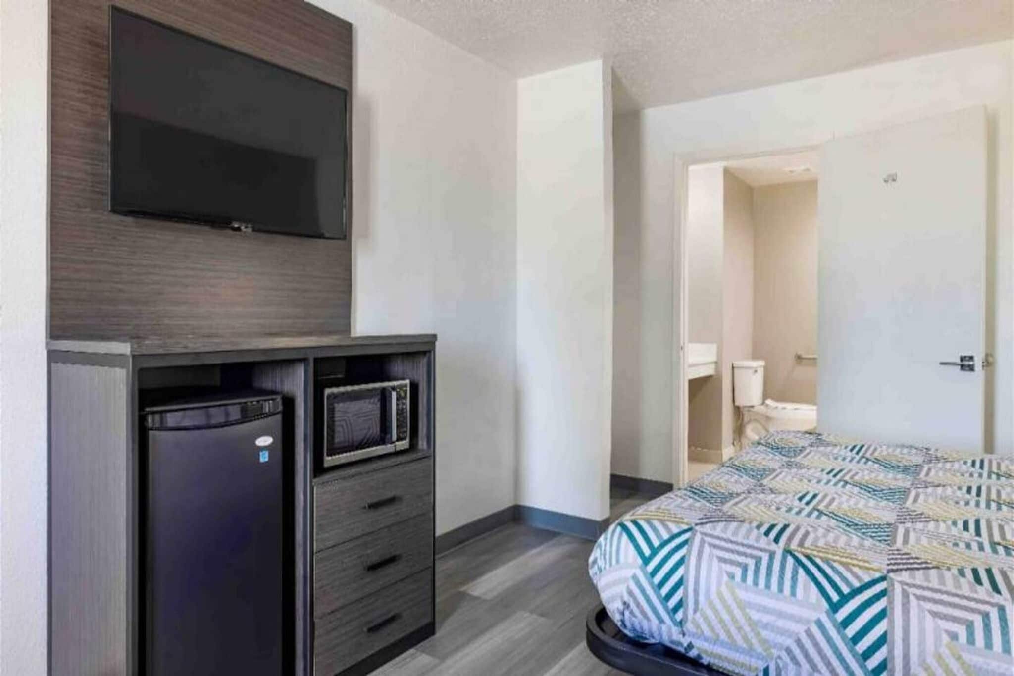 A hotel room with a bed, a TV mounted on the wall, a minibar, and a door leading to a bathroom.