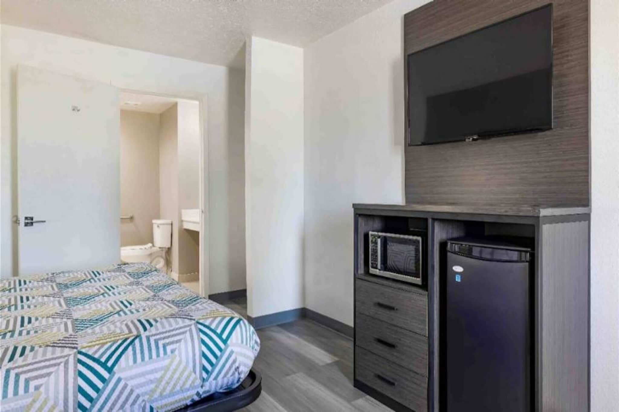 Una habitación de hotel con cama, televisor montado en la pared, escritorio con cajones y puerta que da a otra habitación.