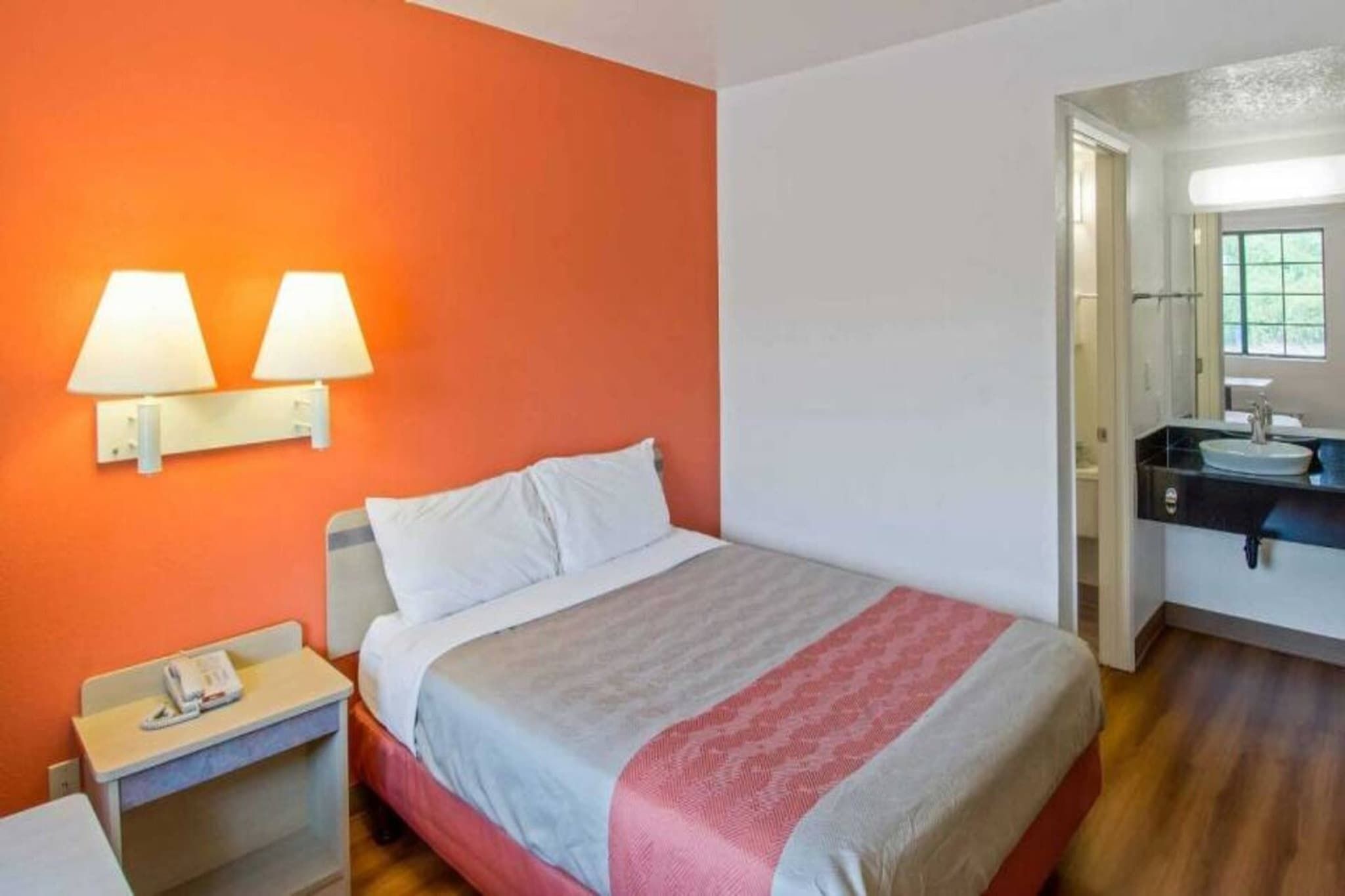 A hotel room with a bed, bedside table, lamp, and a bathroom with a sink and mirror.