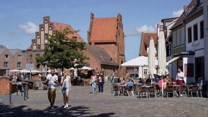 En brostensbelagt gade med udendørs siddepladser, historiske bygninger og en kirke i baggrunden.