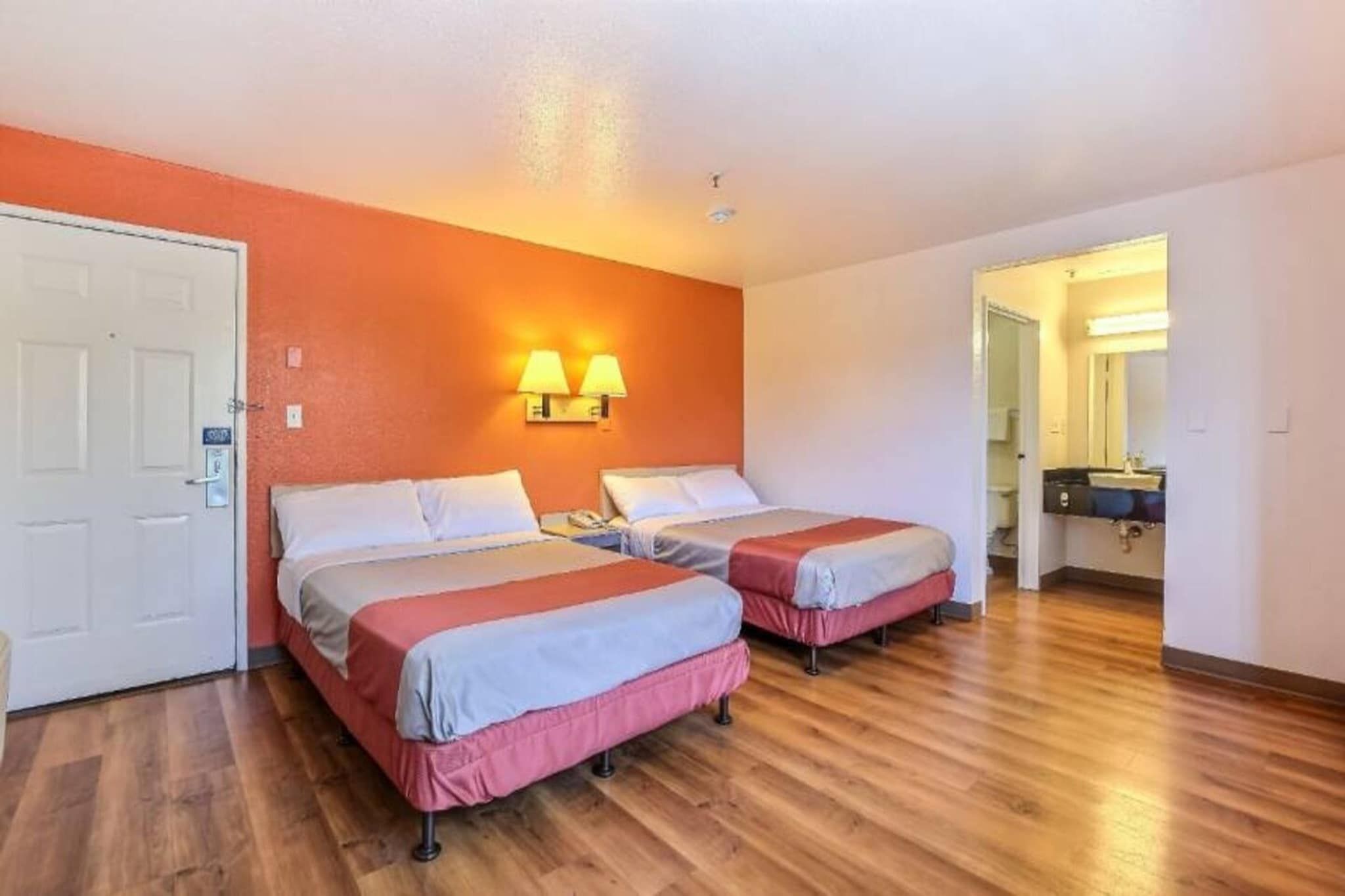 Ein Hotelzimmer mit zwei Betten, Holzboden, einer weißen Tür und einem durch eine offene Tür einsehbaren Badezimmer.