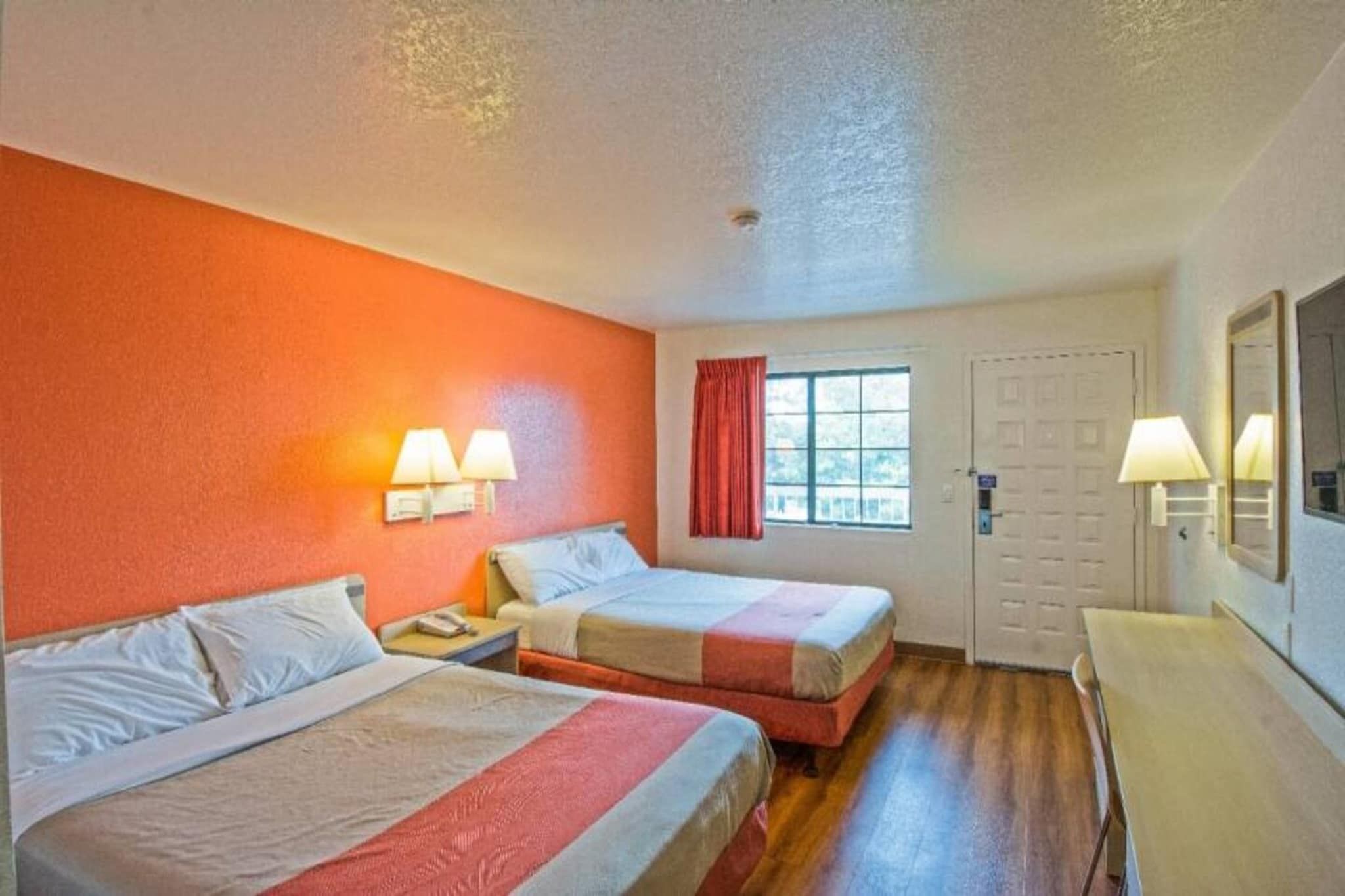 Quarto de hotel com duas camas, uma janela com cortinas vermelhas, uma porta e uma escrivaninha com abajur.