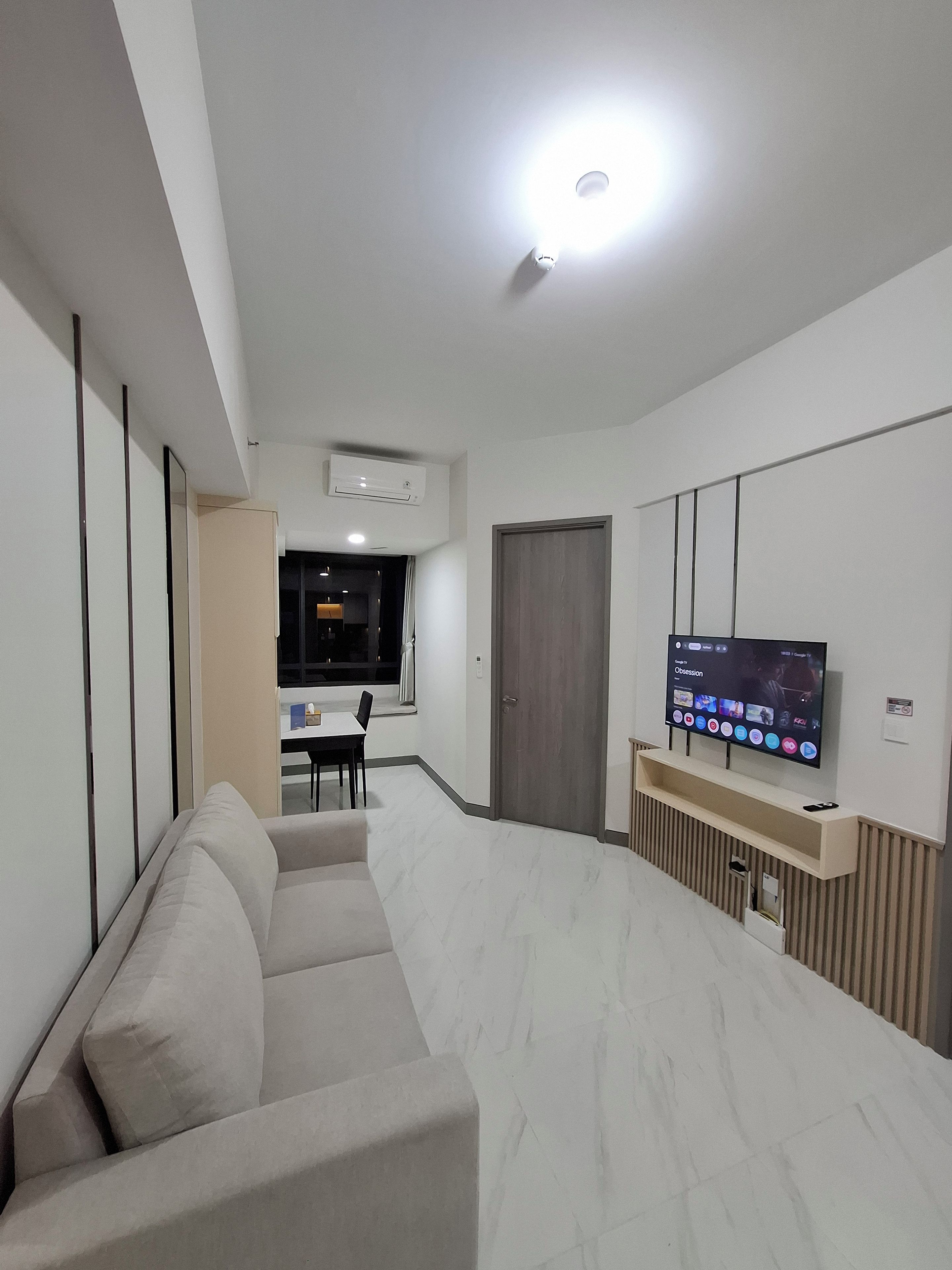 A modern living room with a sofa, a flat-screen TV mounted on the wall, and a dining area in the background.