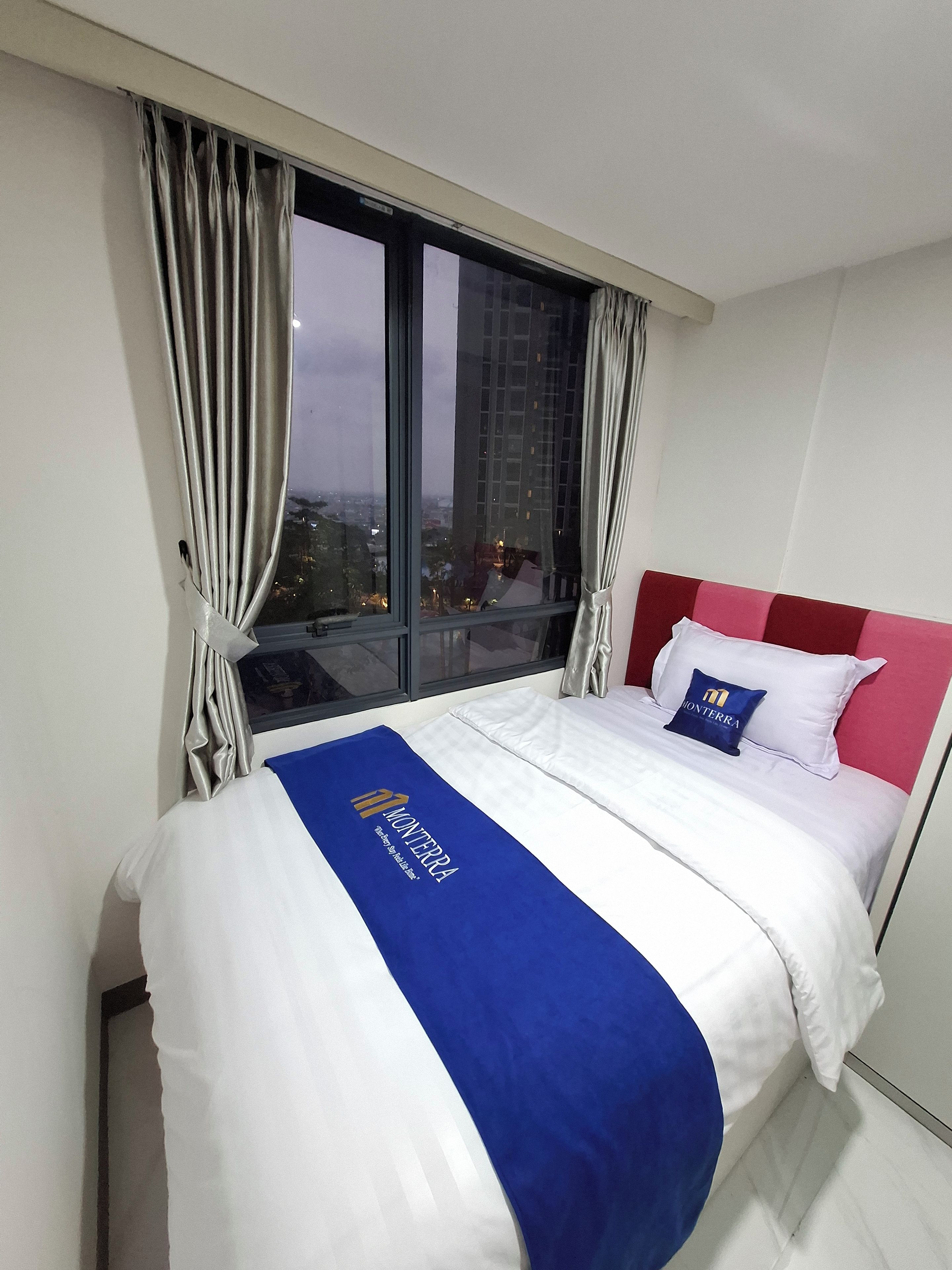 A hotel room with a large bed, a view of the city through a window, and a red headboard.