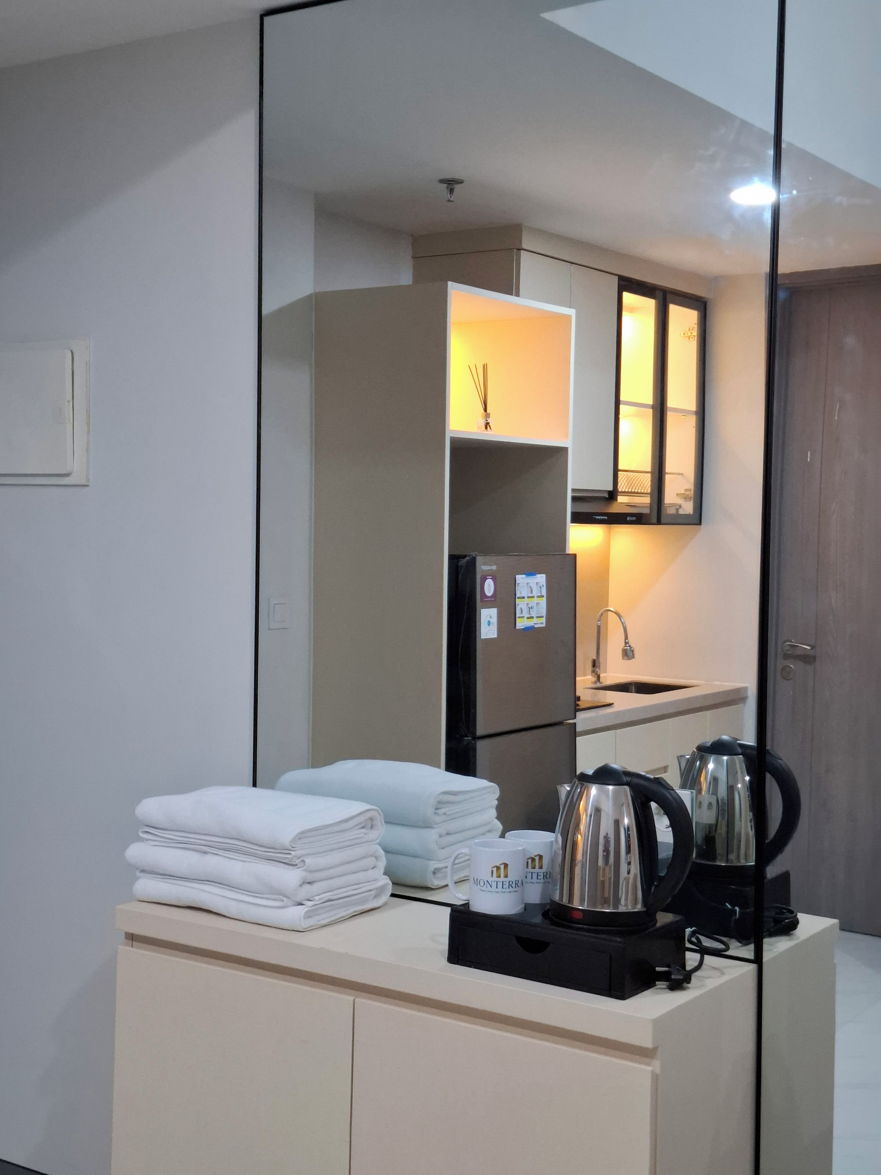 Habitación de hotel con kitchenette, un espejo que refleja el cuarto, un lavamanos y una encimera con toallas y una tetera.