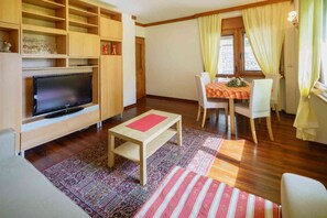 Un soggiorno con televisione, mensole in legno, un tavolo da pranzo e sedie, e un tappeto decorato.