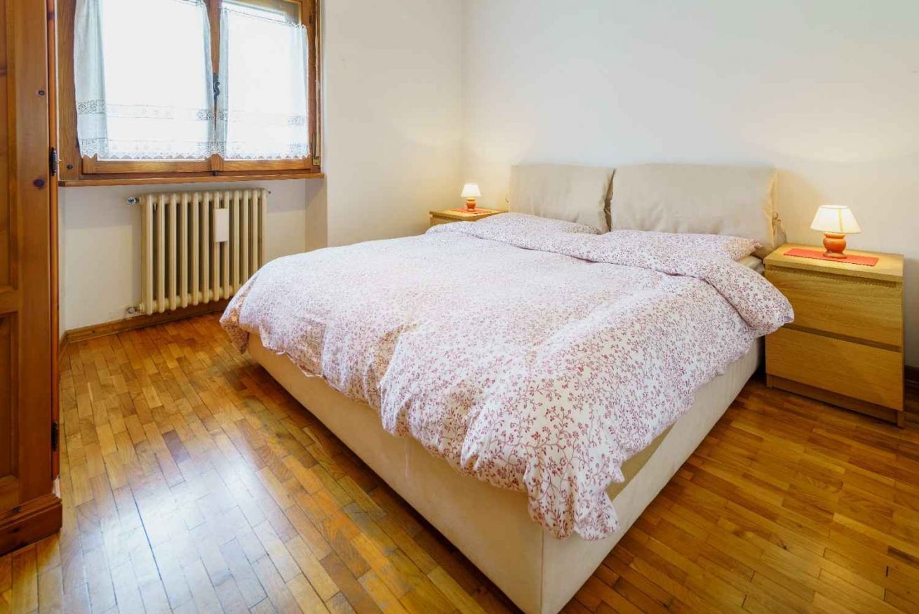 Un dormitorio con suelo de madera, una cama con colcha estampada, dos mesitas de noche con lámparas y una ventana con cortinas.