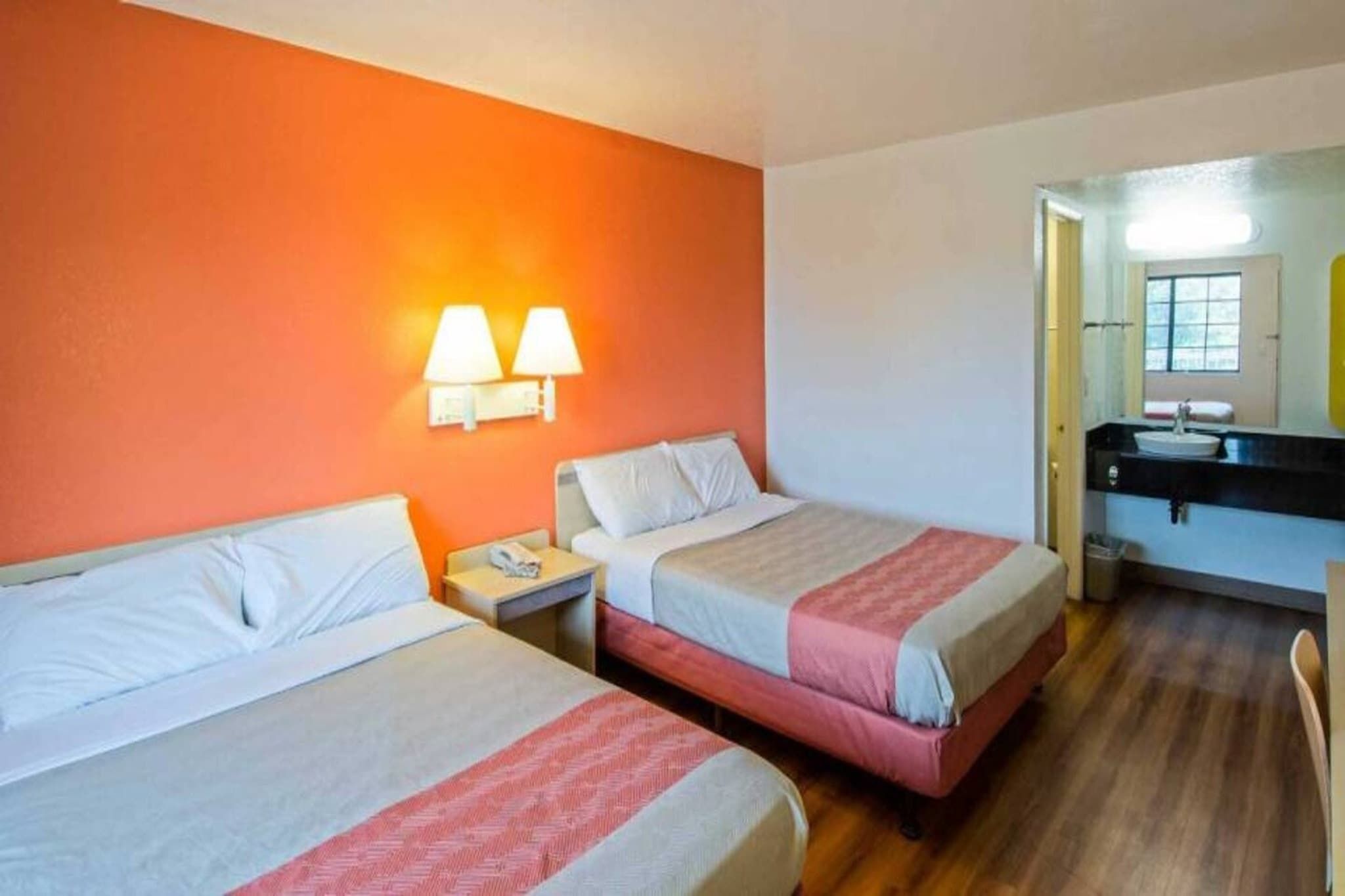 A hotel room with two beds, a nightstand, a bathroom with a sink, and a mirror.