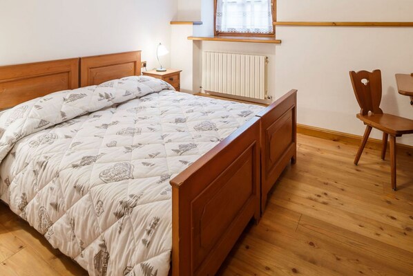 A bedroom with a wooden bed, a bedside table with a lamp, a chair, and a window with a white curtain.