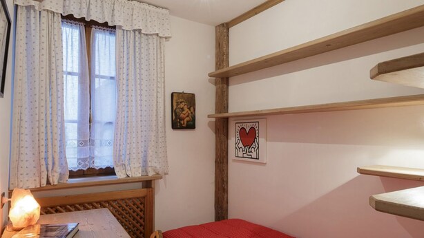 Ein Zimmer mit einem Bett, einem Vorhangfenster, einem Wandregal und einem gerahmten Bild.