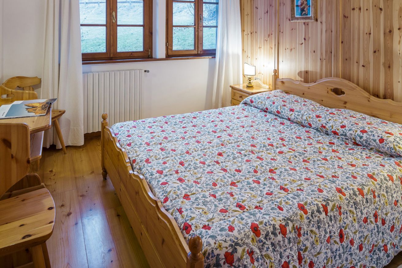 Una cama de madera con una colcha estampada, un escritorio de madera con una silla, una mesita de noche con una lámpara y una ventana con cortinas.