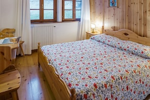 Un llit de fusta amb un llençol floral, un escriptori de fusta amb una cadira, una nitueta amb una làmpada i una finestra amb cortines.