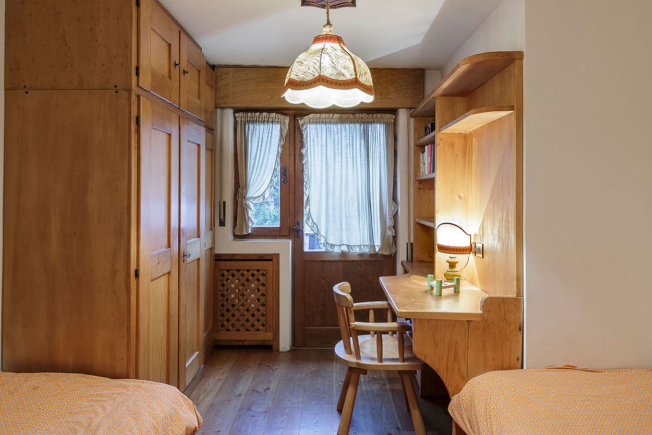 Una habitación con muebles de madera, una ventana con cortinas, una mesa con lámpara y una cama con edredón estampado.