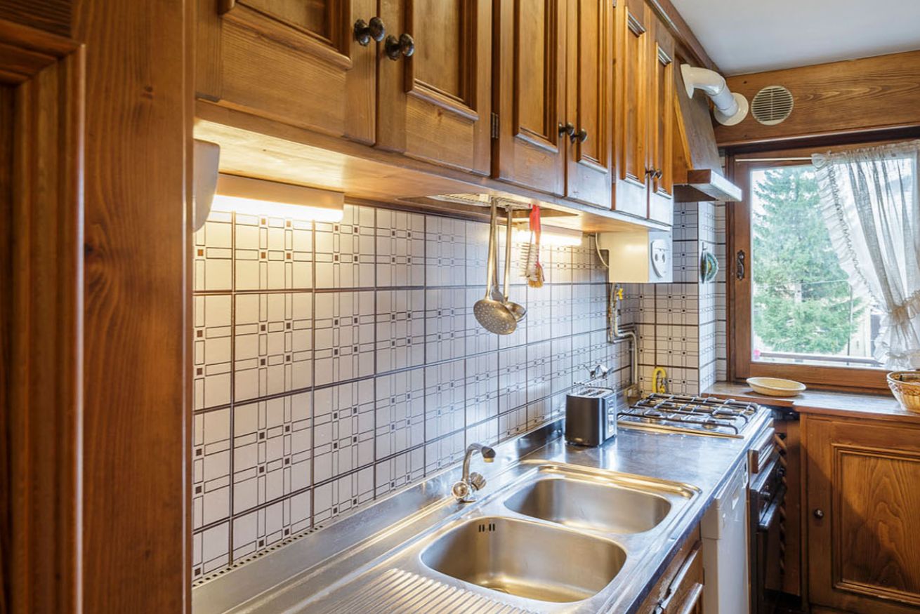 A kitchen with wooden cabinets, a tiled backsplash, stainless steel sinks, and a window with a view of trees.