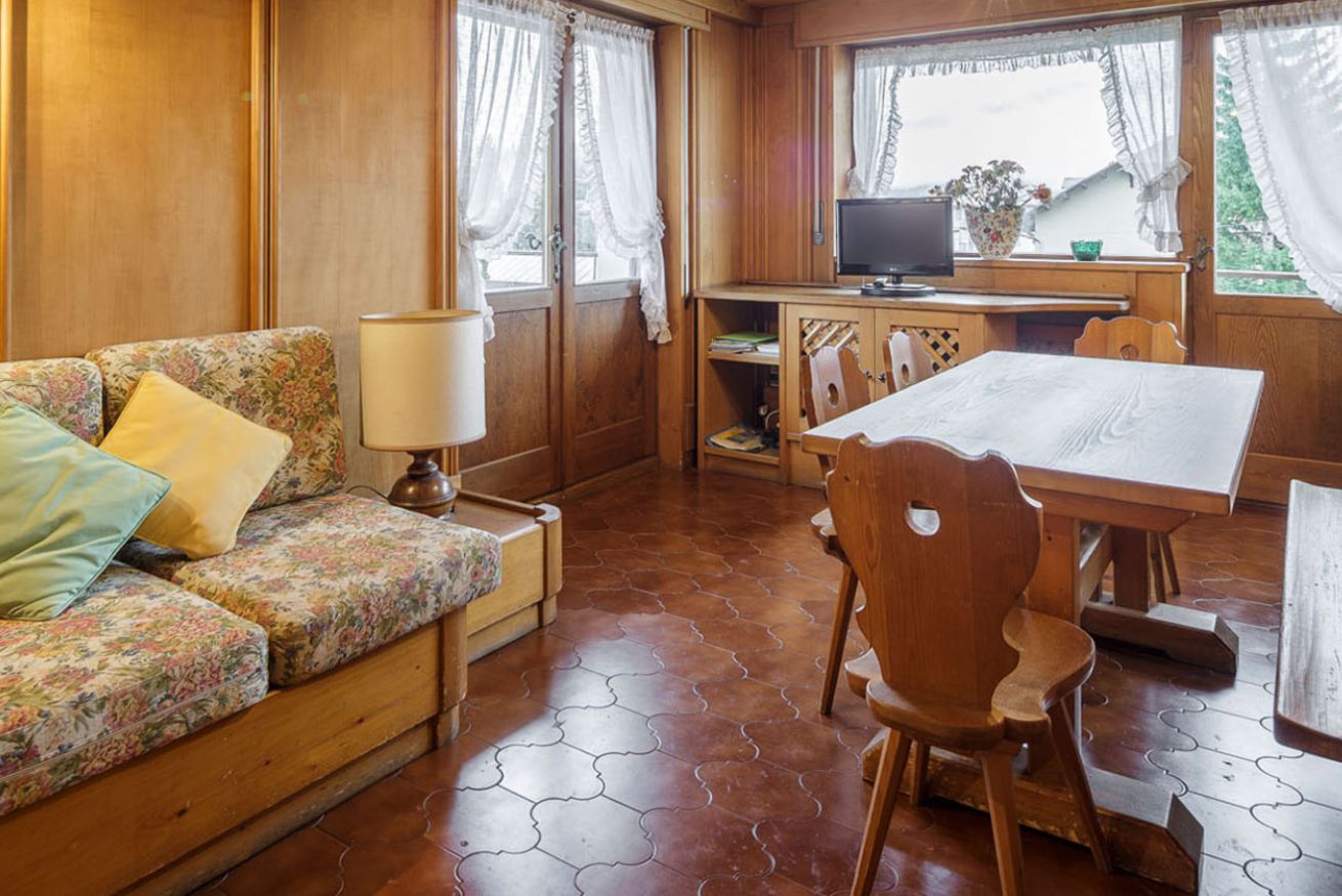 A cozy room with a wooden sofa, a dining table with chairs, a television, and a window with curtains.