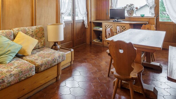 Ein gemütliches Zimmer mit einer Holz-Couch, einem Esstisch mit Stühlen, einem Fernseher und einem Fenster mit Vorhängen.