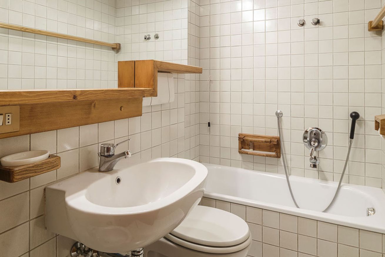 Un baño con lavamanos, inodoro y bañera blancos. Hay repisas de madera sobre el lavamanos y un perchero en la pared.