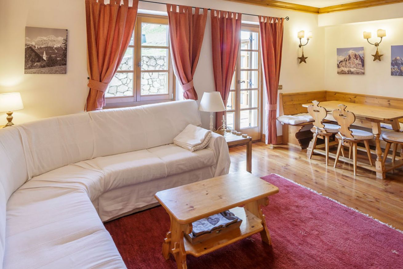 A cozy living room with a sofa, a wooden table, and a bench. There are curtains, a lamp, and wall-mounted lights.