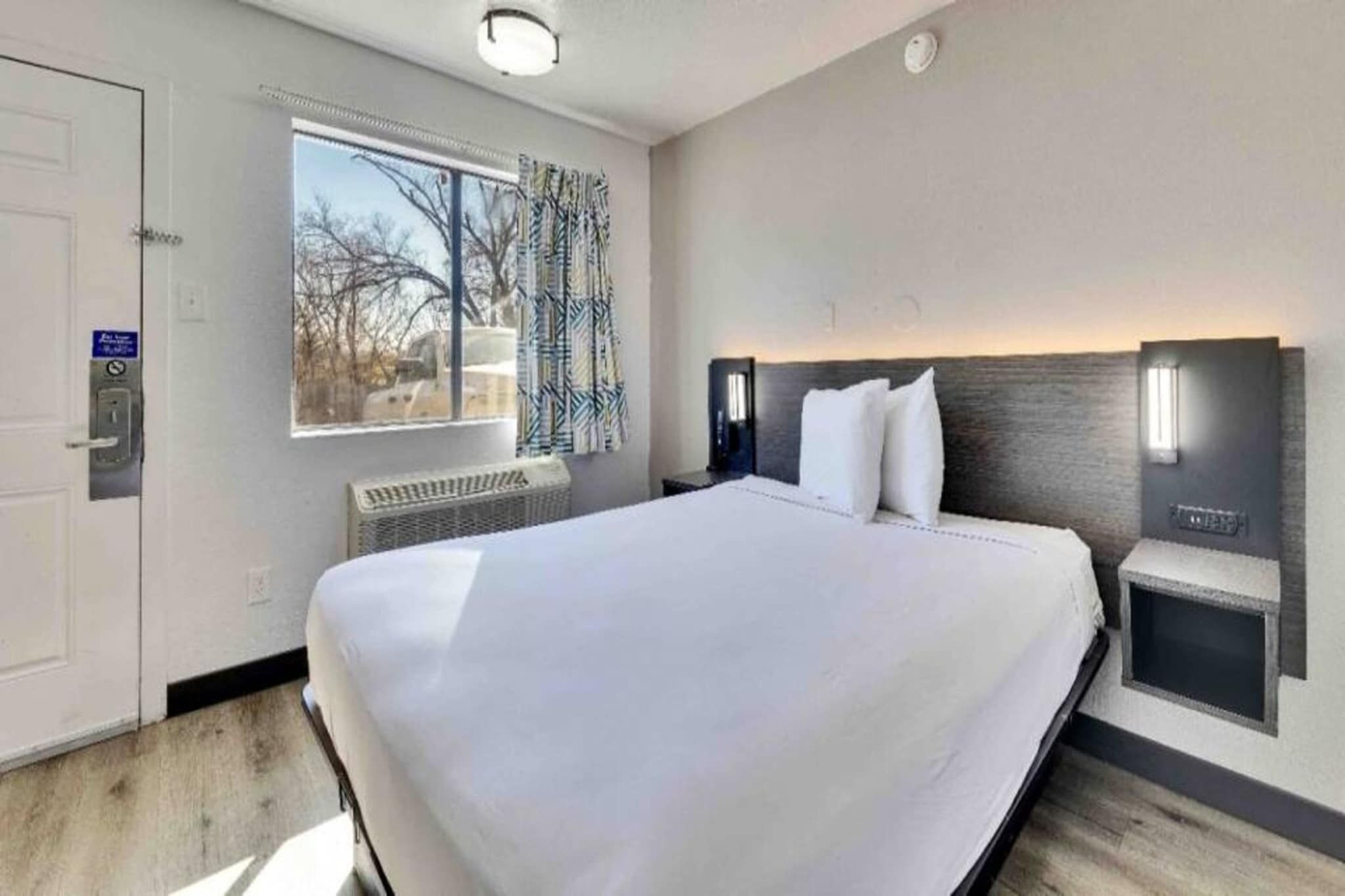 A hotel room with a large bed, a window with curtains, a wall-mounted heater, and a door with a keycard reader.