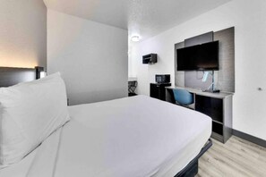 A hotel room with a bed, a desk with a chair, a flat-screen TV mounted on the wall, and a small table with a lamp.