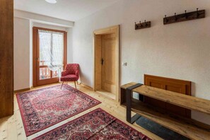Una stanza con una porta in legno, una panchina, un tappeto rosso con decorazioni e una sedia a rombi rossi e bianchi.