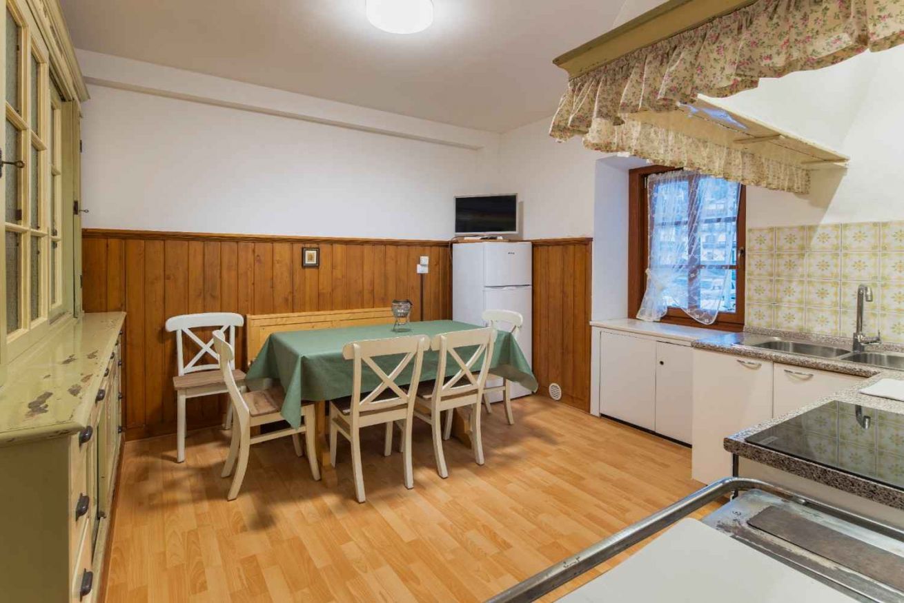 A compact kitchen with wooden cabinets, a dining table with chairs, a television, and a window with a curtain.