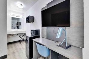 A modern hotel room with a flat-screen TV mounted on the wall, a desk with a chair, and a small kitchenette area.