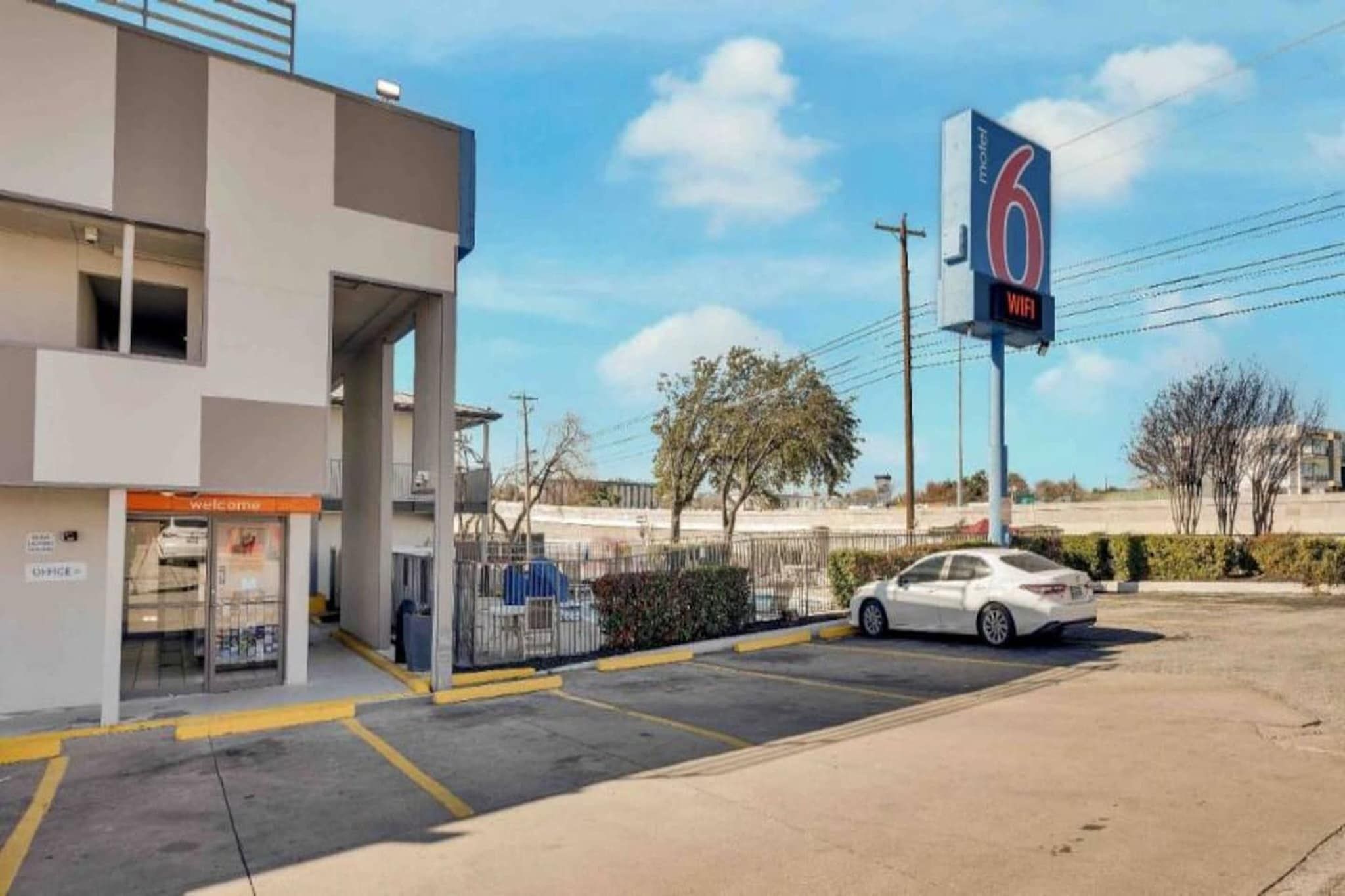 A motel with a large sign indicating the number 6. There is a car parked in the lot, and a building with a 'Welcome' sign.