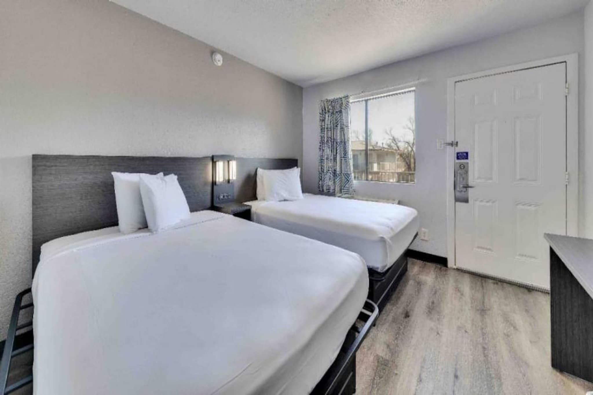 A hotel room with two beds, a window with patterned curtains, a door with a lock, and a small desk.