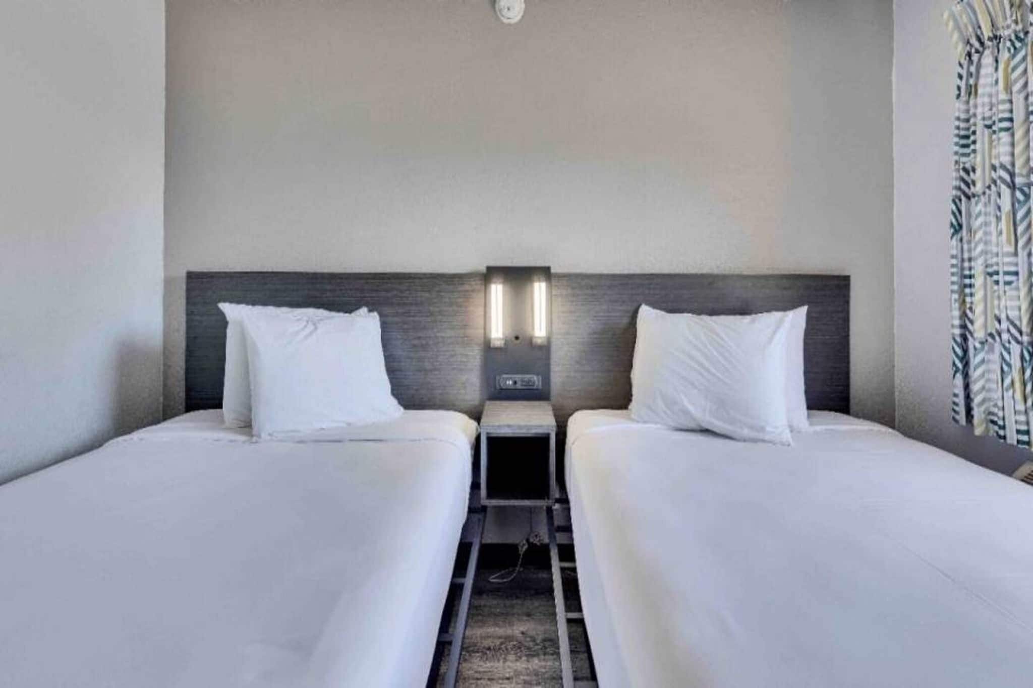 A hotel room with two beds, a nightstand, and a wall-mounted light fixture.