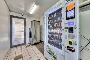 En automattankstation med en digital display och en QR-kod, placerad i en korridor med kakelgolv och en dörr.