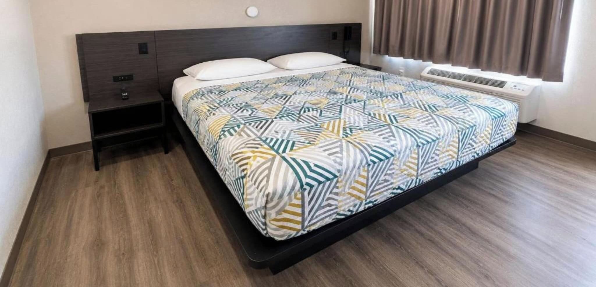 A hotel room with a bed, a nightstand, a heater, and a window with curtains.
