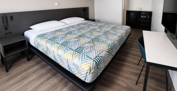 A modern hotel room with a bed, bedside table, chair, and kitchenette.