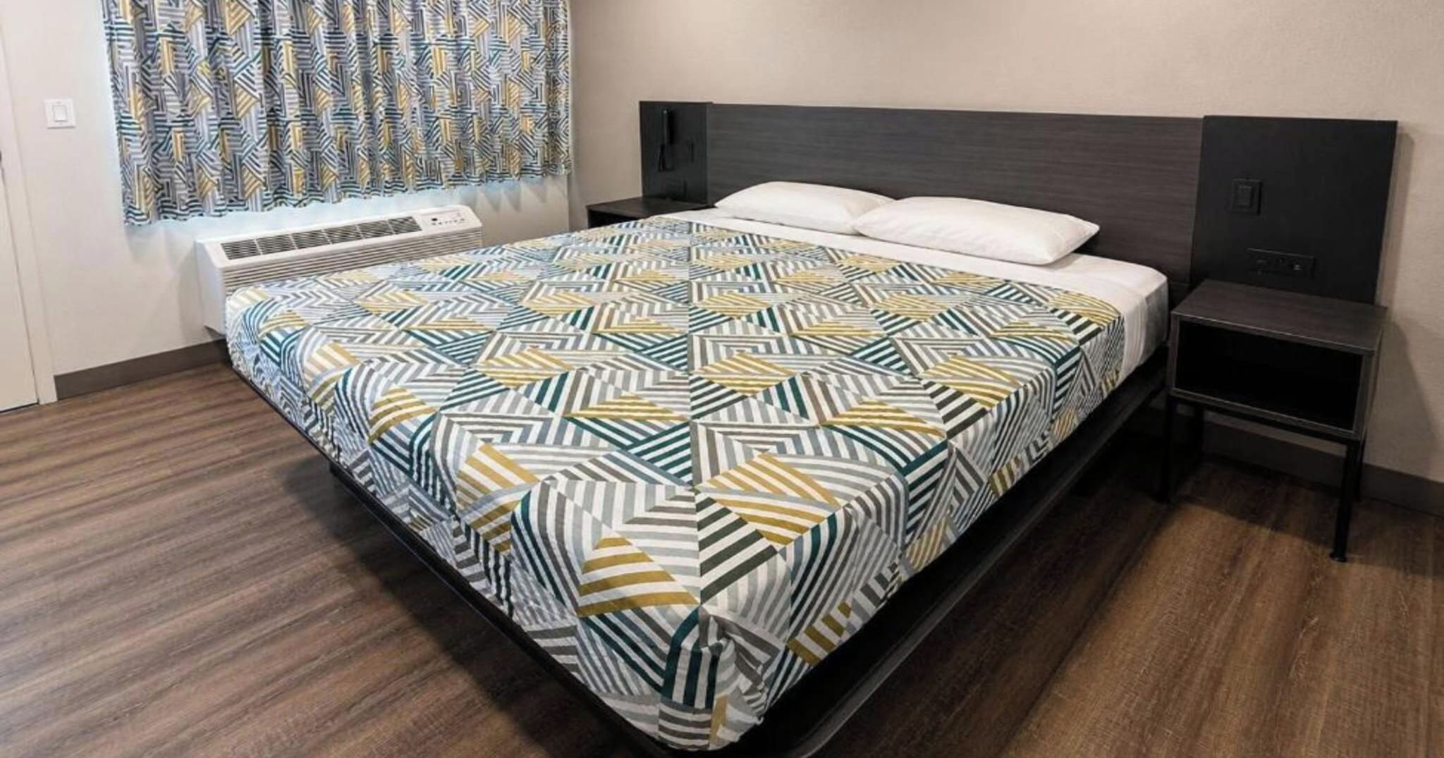 A hotel room with a bed, a nightstand, a wall-mounted heater, and a window with patterned curtains.