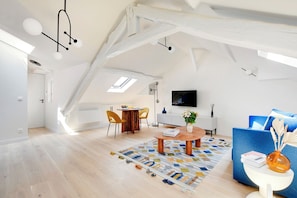 Un loft espaiós amb sostres alts, bigues descobertes, zona de menjador amb taula rodona, televisió penjada a la paret i un sofà blau.