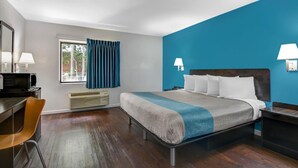 A hotel room with a bed, a desk, a chair, a window with curtains, and a wall-mounted lamp. - 1 King Bed | Non-Smoking (Norcross)