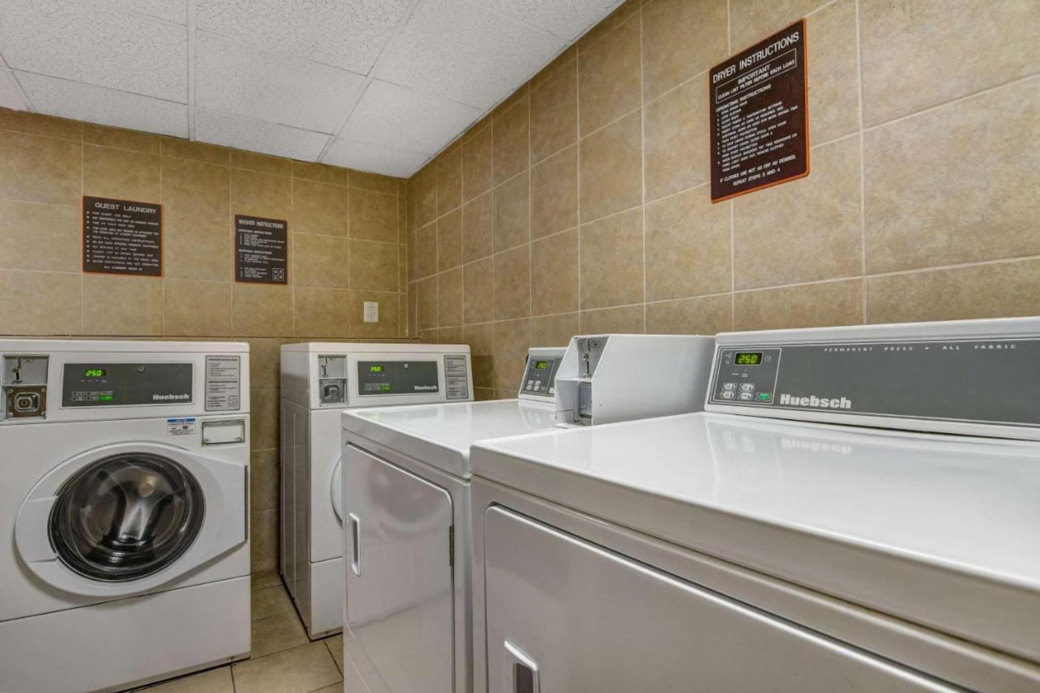 A laundry facility with multiple machines, including a front-loading washer and a top-loading dryer, with instructions and labels on the wall.